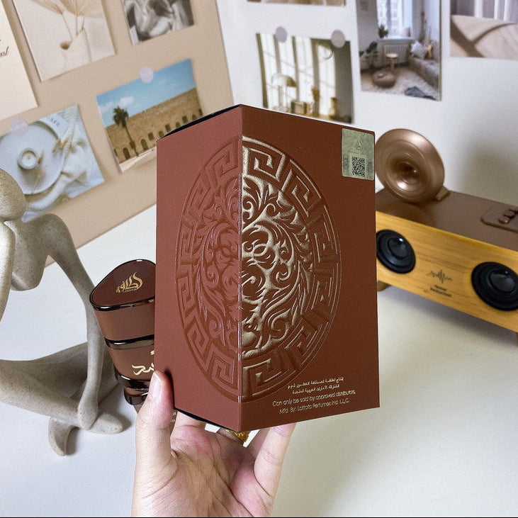 A hand holds a brown rectangular product box with embossed circular ornamental detailing. The box is photographed in a styled indoor setting with a speaker, figurine, and framed wall art in the background.